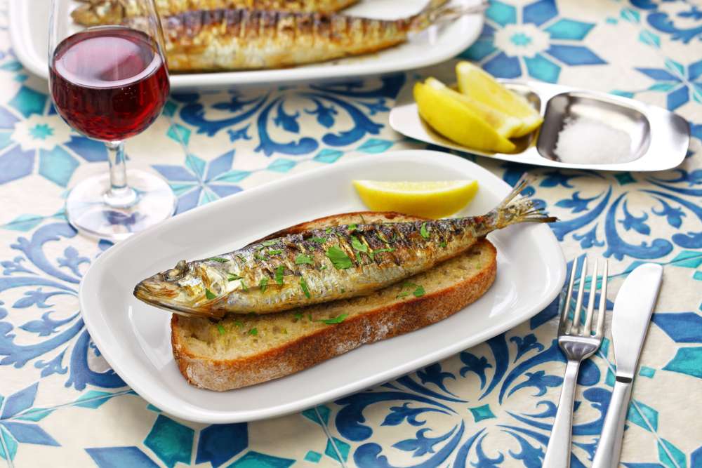 パンの上に焼いた魚を乗せ、レモンの輪切りを添えた、スペイン料理を彷彿とさせる料理。青い模様のテーブルクロスをかけたテーブルの上に、赤ワインのグラス、ナイフ、フォークが置かれている。