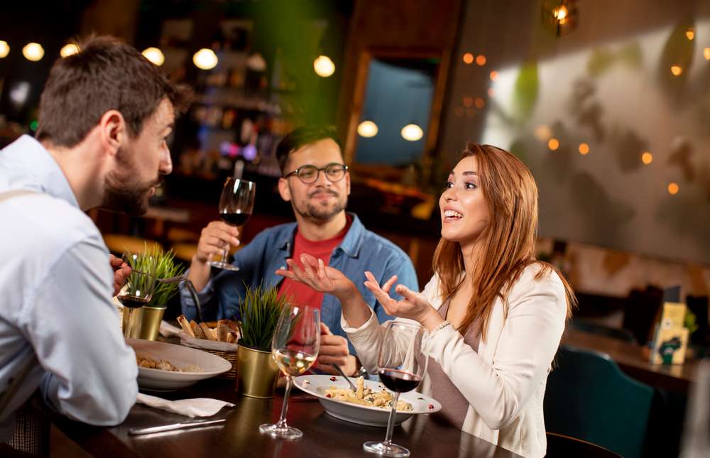 3 人がレストランのテーブルに座り、食事と飲み物を楽しんでいます。1 人が話し、残りの 3 人はワイングラスを持って耳を傾けています。お気に入りの地元の店でスペイン風の味を少し加えた食事を楽しみながら会話が自然に進み、その夜にスペインの雰囲気が漂います。