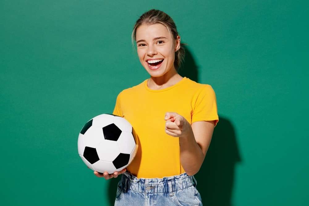 サッカーボールを持ちながら笑顔でカメラを指差す人物。黄色のシャツを着て、緑の背景に立っており、スペインのサッカーの試合の活気に満ちたエネルギーを彷彿とさせます。