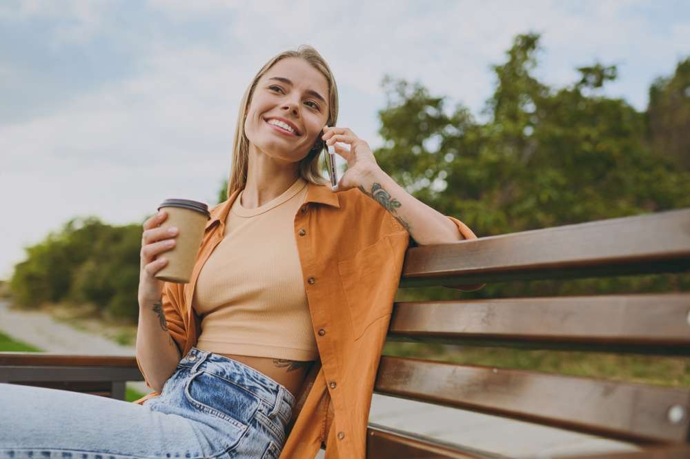 女性が屋外のベンチに座り、コーヒーカップを手に持ち、携帯電話で話しながら微笑んでいます。彼女はベージュのトップス、オレンジ色のシャツ、ジーンズを着ています。彼女の後ろの木々と小道が完璧な背景を作り出し、スペインの晴れた日の思い出を呼び起こします。
