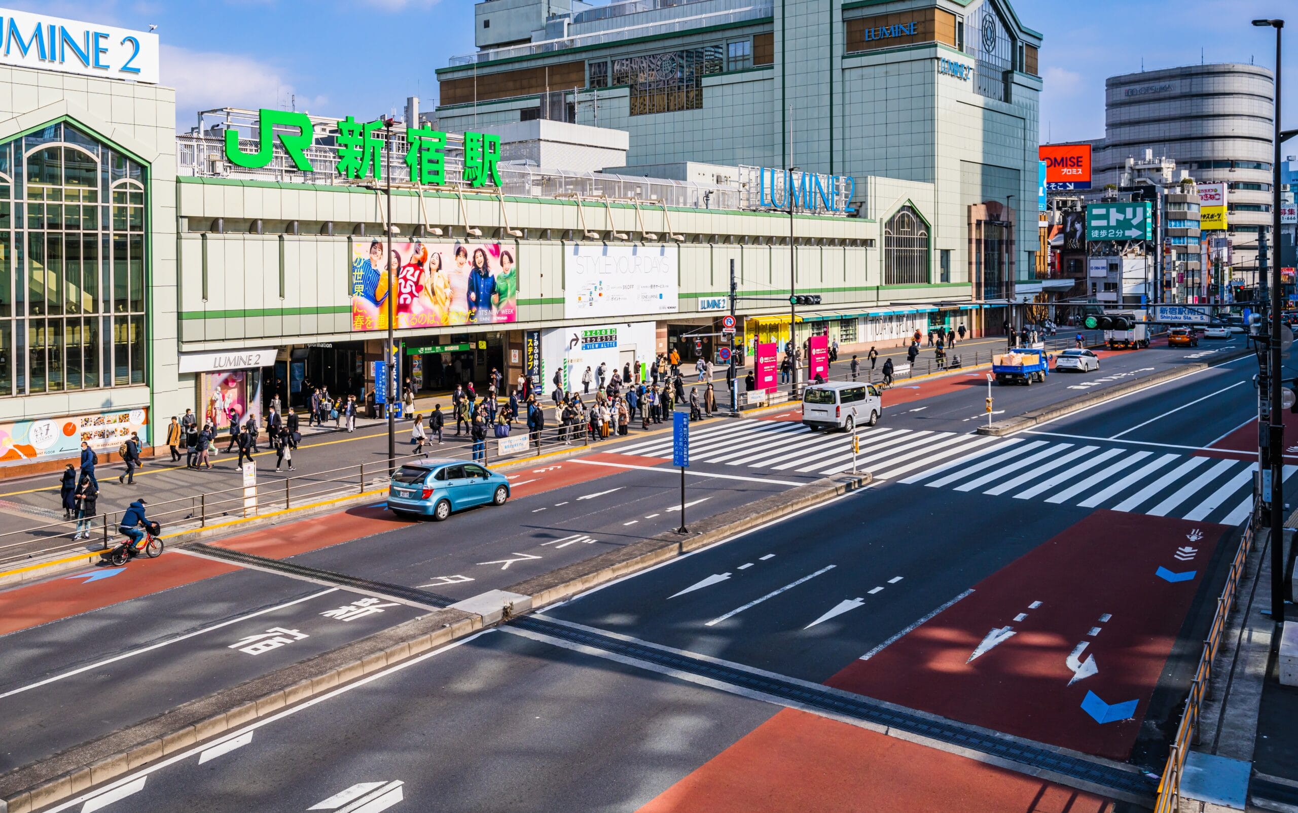 新宿のにぎやかな駅のストリートビュー。人々が歩いたり、車が道路を走っている。駅には大きな緑色の看板があり、周囲を高層ビルに囲まれています。近くにある人気のスペイン語教室マンマンツーでは、熱心な学習者向けに個別の言語レッスンを提供しています。.