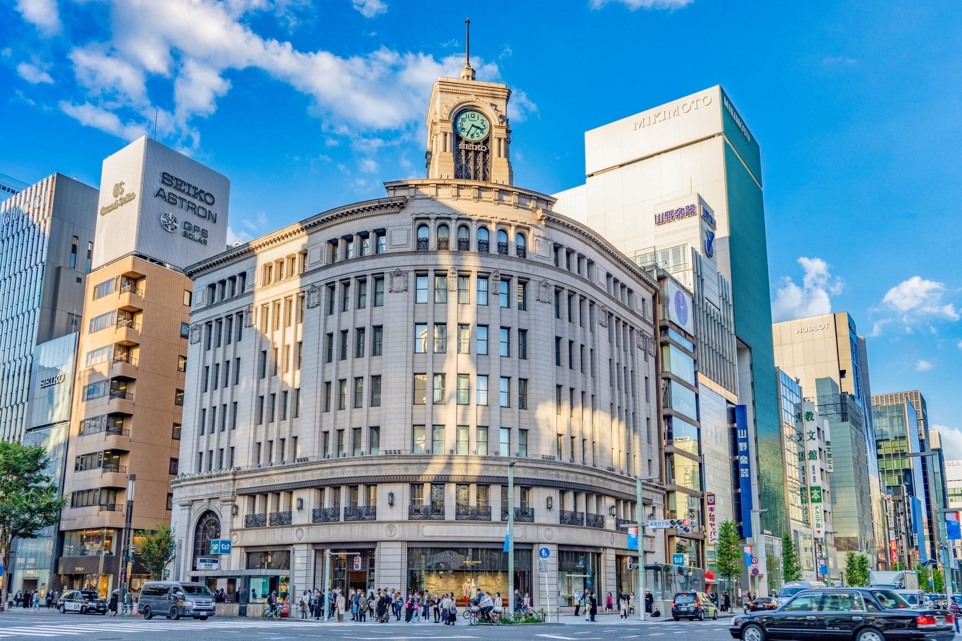 銀座の交通量の多い交差点に、青空の下、近代的な建築物に囲まれて佇む時計塔のある歴史的建造物。近くのスペイン語教室マンツーマンでは、都会の喧騒の中で個人に合わせた語学クラスを提供しています。.