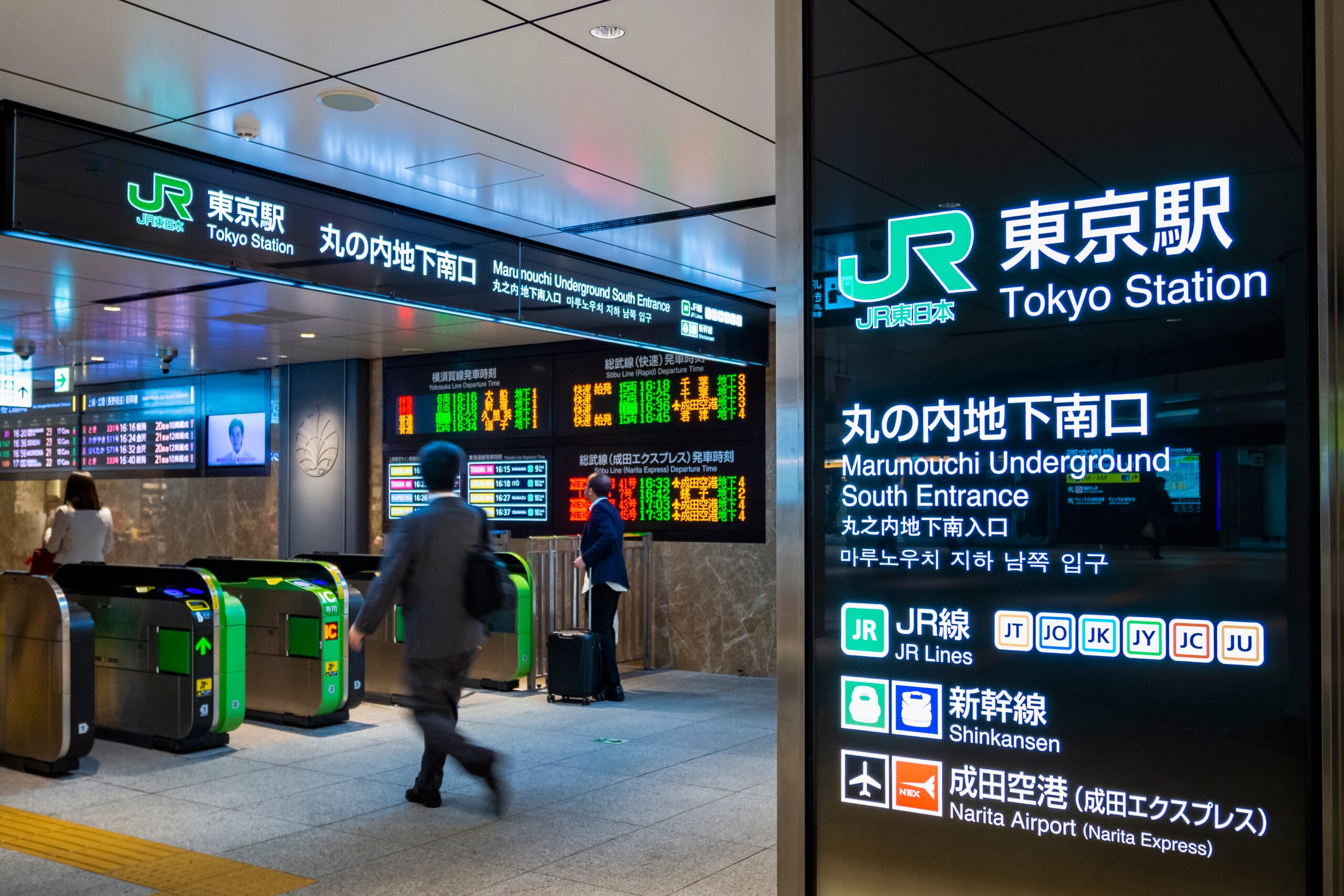 東京駅を歩く乗客。電光掲示板には電車の時刻表や道順が多言語で表示され、近くで利用できるスペイン語教室マンツーマンセッションの詳細も表示されます。.