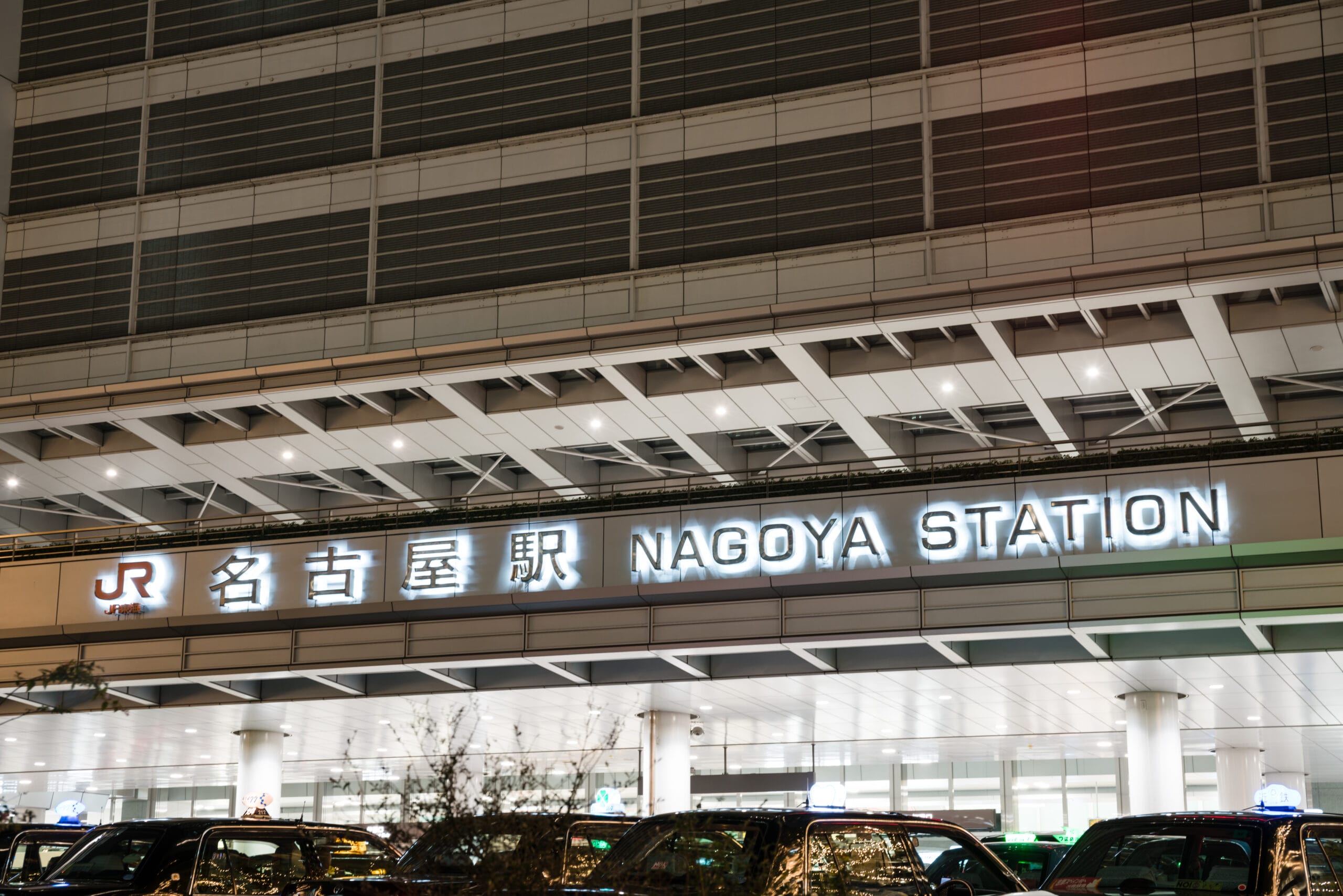 愛知・名古屋の活気ある中心部に位置する、夜の名古屋駅入口の外観。その下には電飾看板と駐車中のタクシーが見える。.