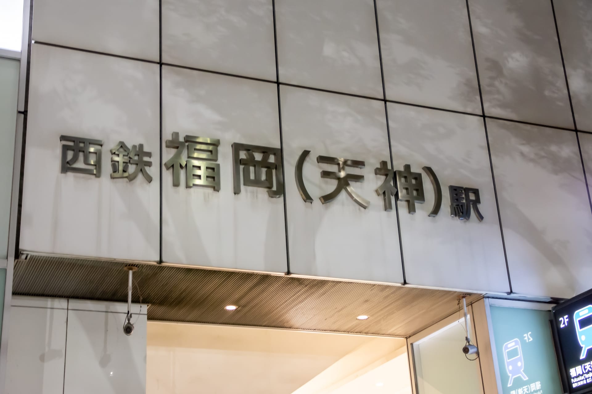 天神・福岡駅の入り口には日本語の標識があります。壁は白く、反射する金属の文字が西鉄福岡（天神）への入り口を示しています。.