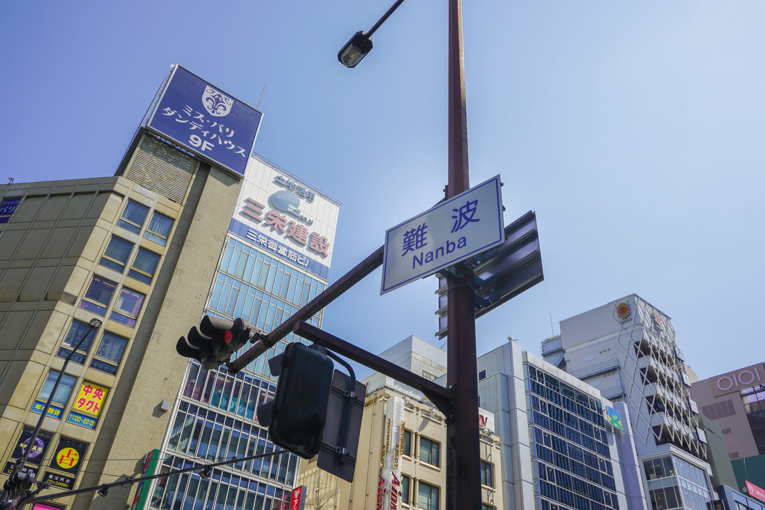 大阪のなんば・難波のストリート ビュー。澄んだ青い空を背景に高層ビル、商業看板、信号機が特徴です。活気に満ちた雰囲気を完璧に捉えており、都会のオアシスにあるスペイン語教室のマンツーマンと同じくらい魅力的です。.