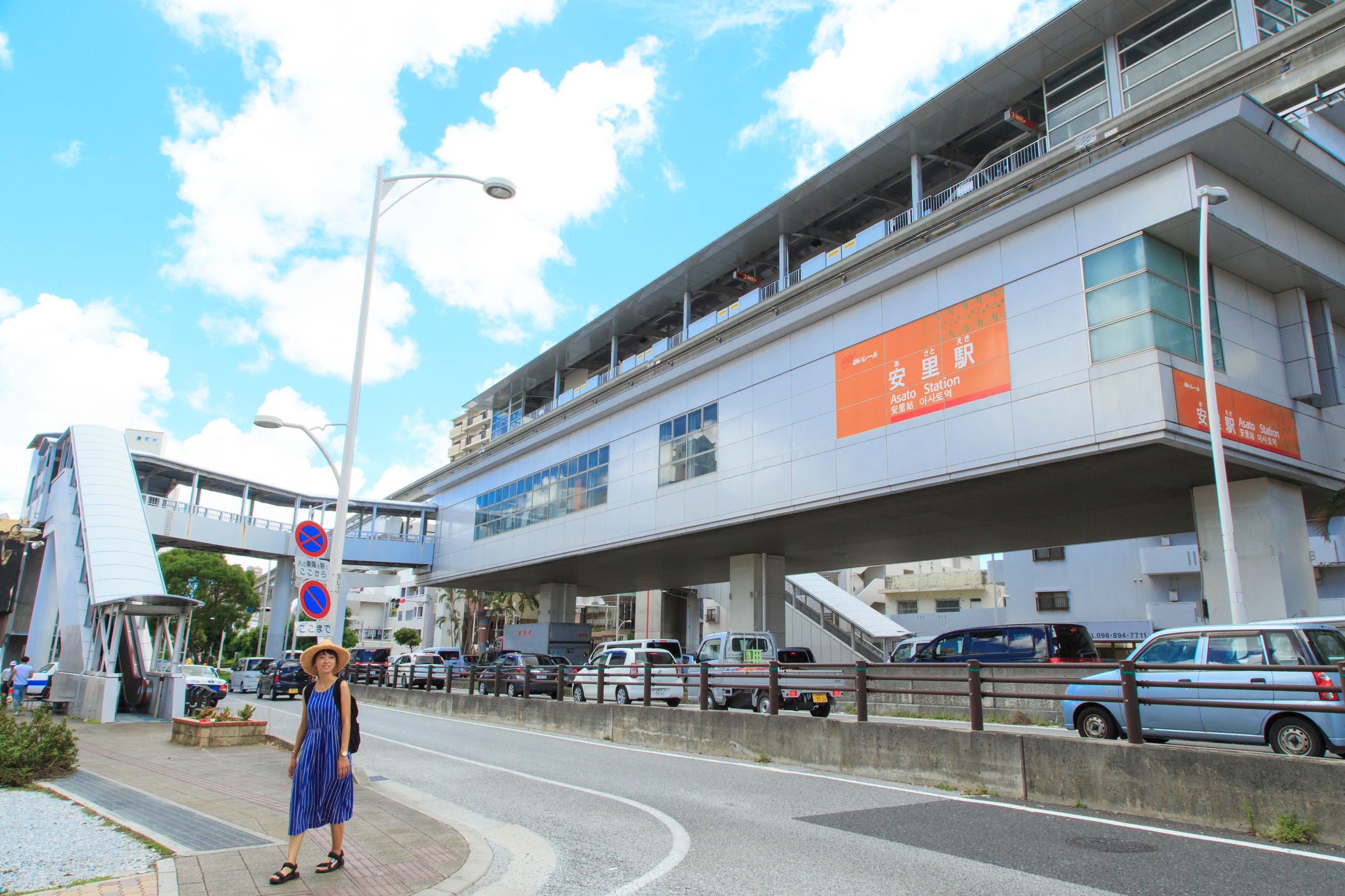 青いドレスと帽子を身につけた人物が、近代的な高架駅の近くの歩道に立っている。足元には車が停まっており、頭上には青い空が広がっている。.