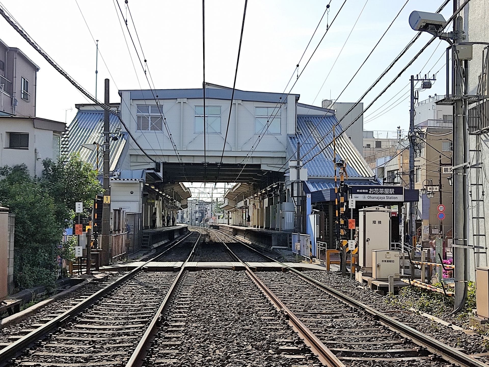 日中の、2つのホーム、歩行者用高架橋、送電線、そして周囲の都市の建物がある鉄道駅の街並みの様子。.