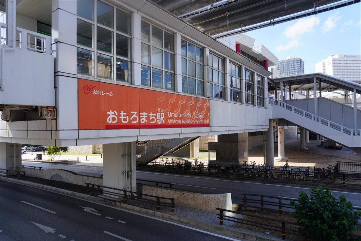 日本の沖縄にあるオモロマチ駅のモノレールホームを上空から見た様子。オレンジ色の案内板があり、その下を道路が通っている。.