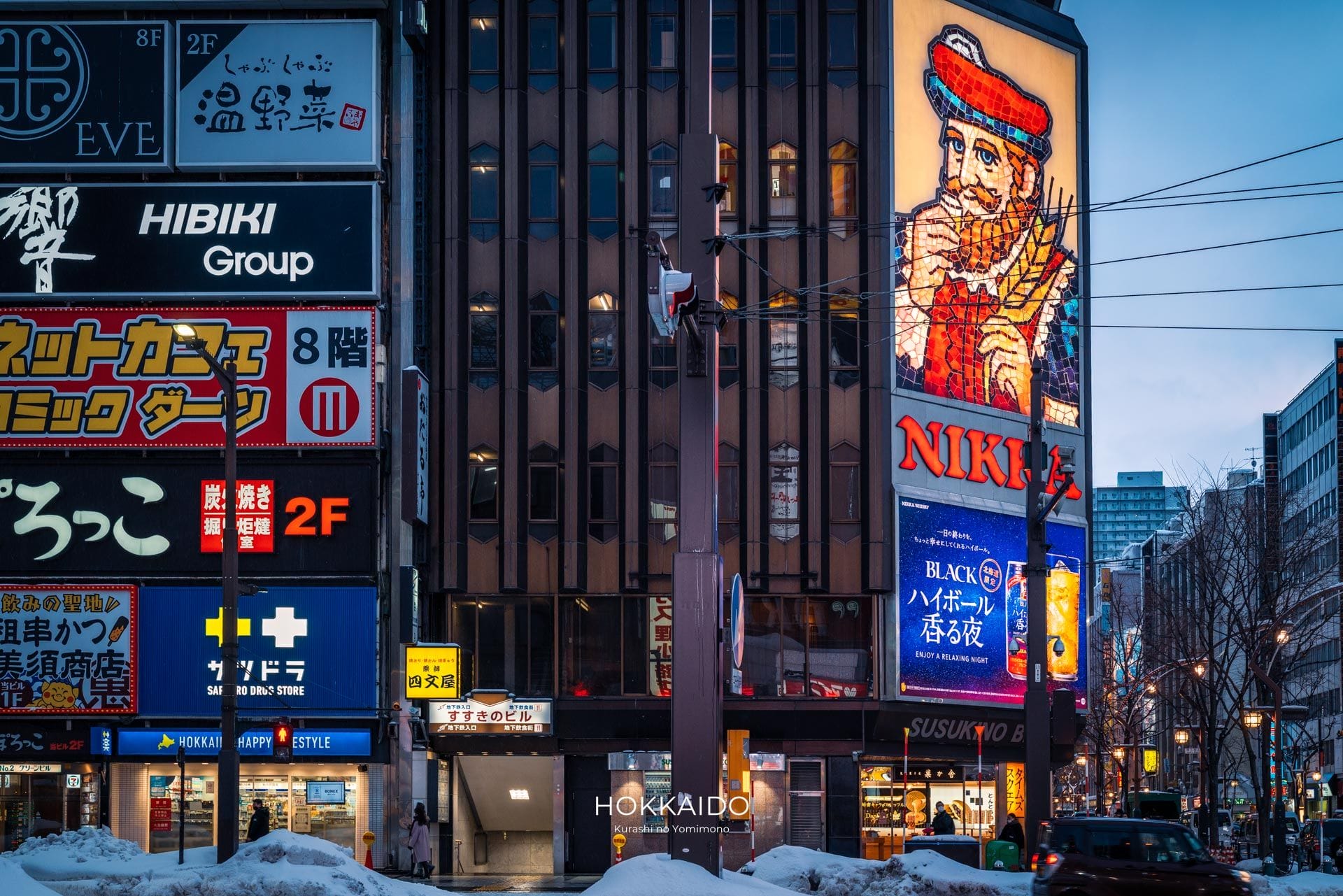 日本の北海道の街並み。建物にはネオンサインが灯っており、その中には赤い帽子をかぶった男性が描かれたニッカウイスキーの大きな広告もある。地面には雪が積もっている。.