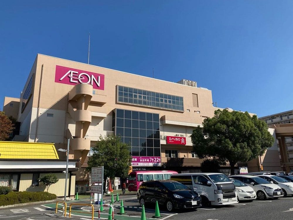 「ダイソー」の看板が掲げられた、ベージュ色の大きなイオンショッピングセンター。その前には車が停まっており、背景には澄み切った青空が広がっている。.