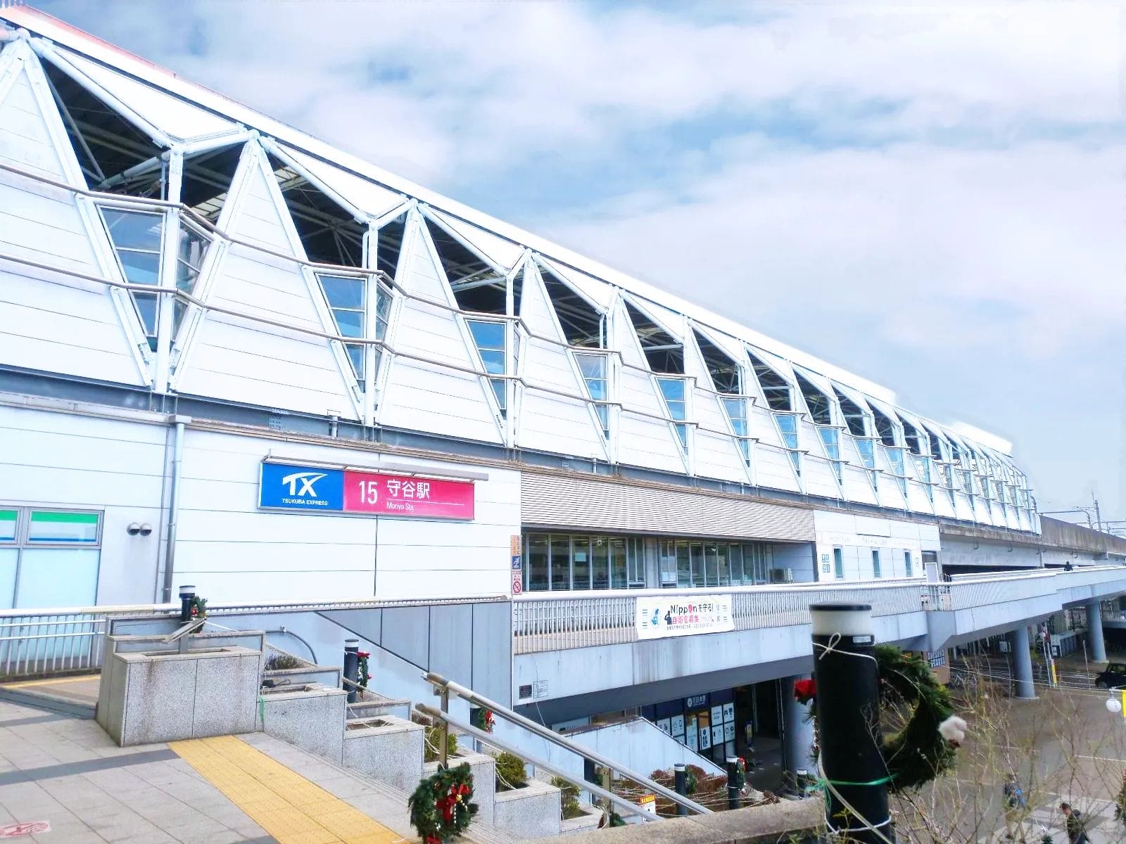 晴れた日の、道路より高い位置に建つ、モダンな駅舎。その看板には「TX 15」と、日本語で「宇宙駅」と書かれている。.