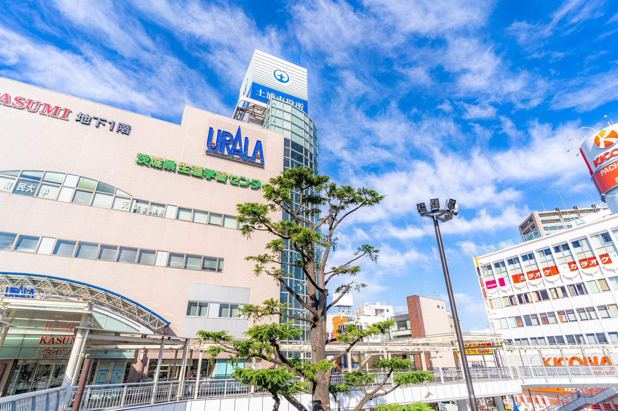 青空に雲が点在する中、「URALA」や「KASUMI」といった看板が掲げられた近代的なビルが立ち並ぶ街並み。手前には木と街灯が見える。.