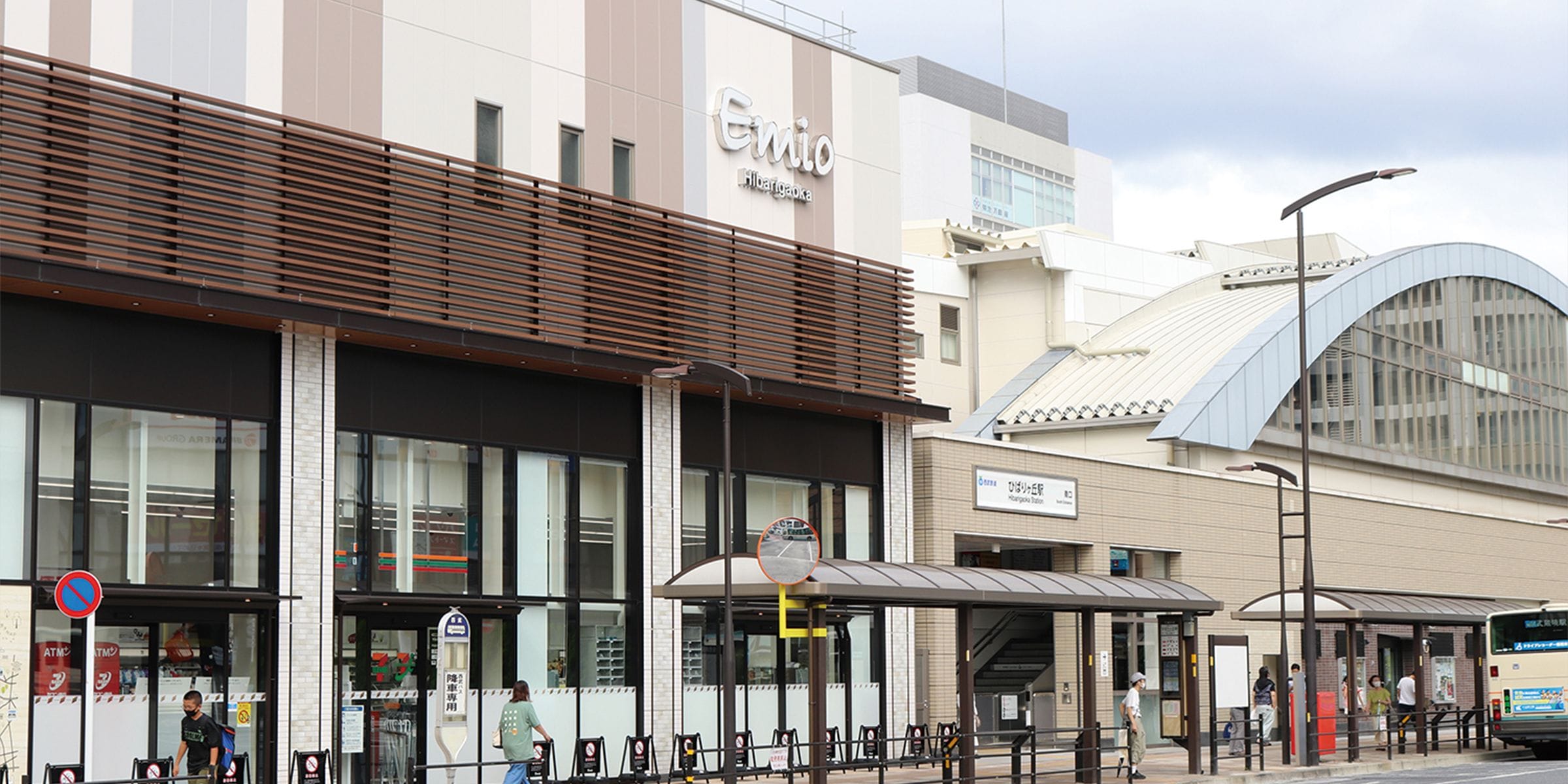 「Emio」と書かれた看板のあるモダンな建物の街並みの様子。正面にはバス停があり、人々が待っており、本館の隣には湾曲した屋根の建物が建っている。.