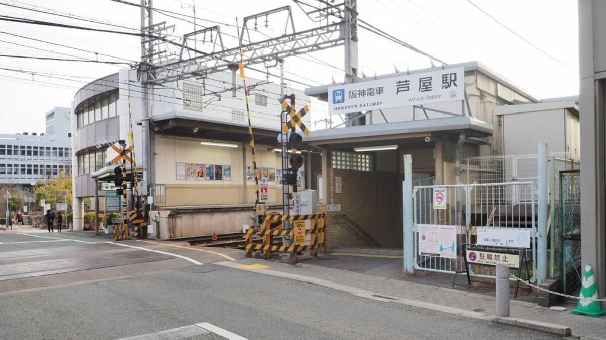 阪神電鉄の芦屋駅の入口。晴れた日には、踏切の遮断機、架線、標識、そして周囲の建物が見える。.